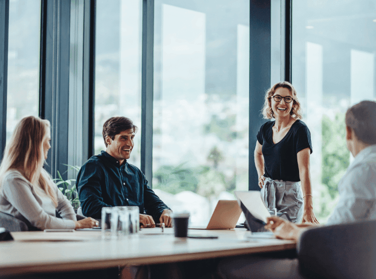 Four business professionals in a modern office meeting room with floor-to-ceiling windows and city views in background.