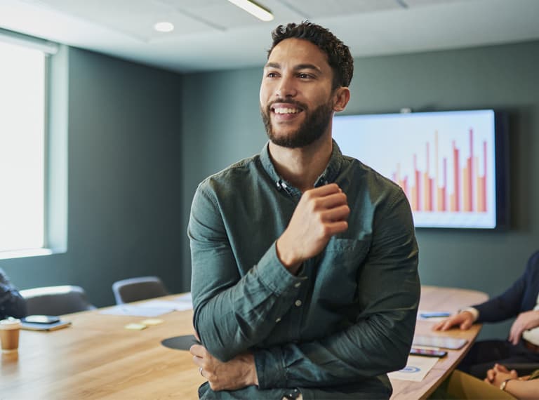 Confident young businessman standing in a modern office and smiling. He is wearing a casual shirt and has a beard. In the background, there are people sitting at a conference table.