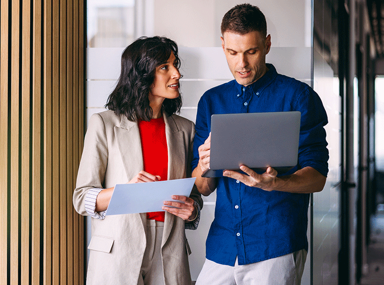 Two business professionals reviewing documents together, woman in beige blazer and red top, man in blue shirt holding laptop.