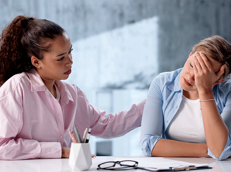 Woman in pink shirt comforting distressed colleague in blue shirt at office desk with glasses and clipboard visible.