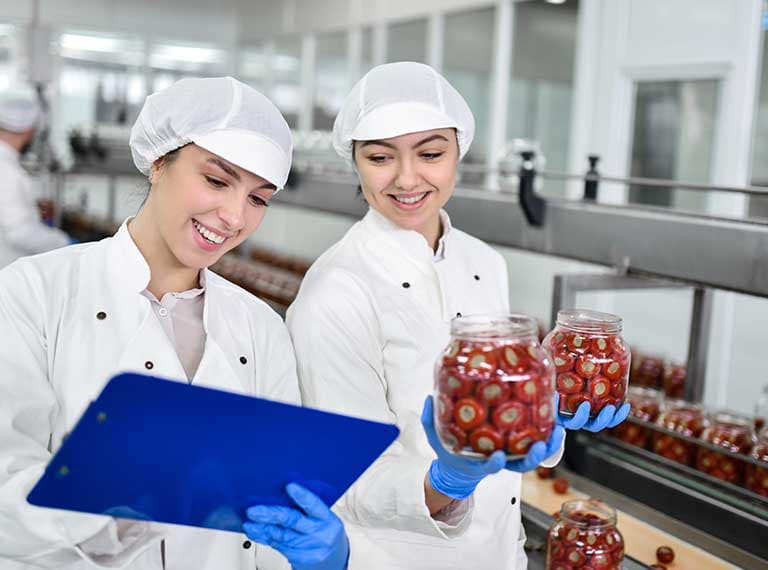 Two food production workers in white uniforms and hairnets examining jars of preserved red vegetables in a factory setting.