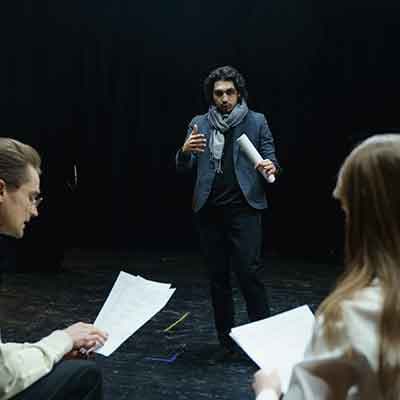 Actor with curly hair in gray blazer gesturing expressively on dark stage while two people with scripts watch from foreground