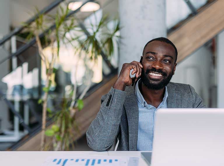 Person in gray blazer smiling while talking on phone in modern office with plants and staircase in background.