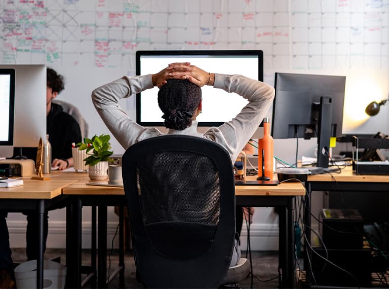 Rear view wide shot of a woman sitting working at a computer in an office. She has her hands behind her head as she stretches, looking at the screen.