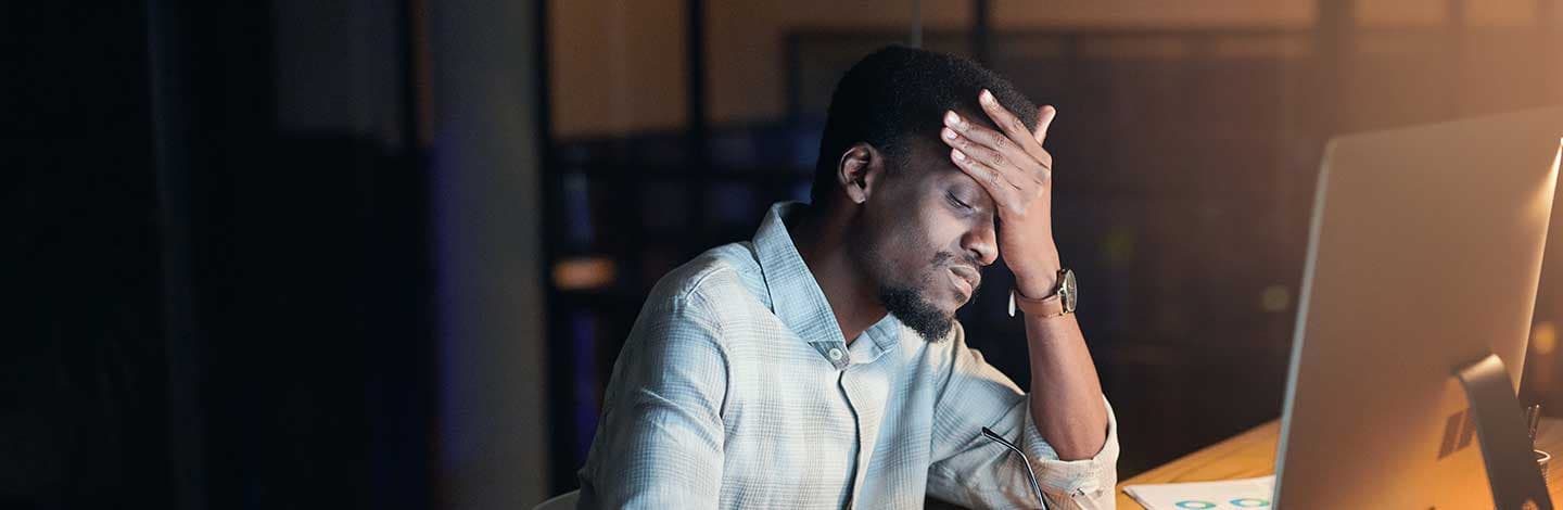 Stressed man in light blue shirt with hand on forehead working late at computer in dimly lit office.
