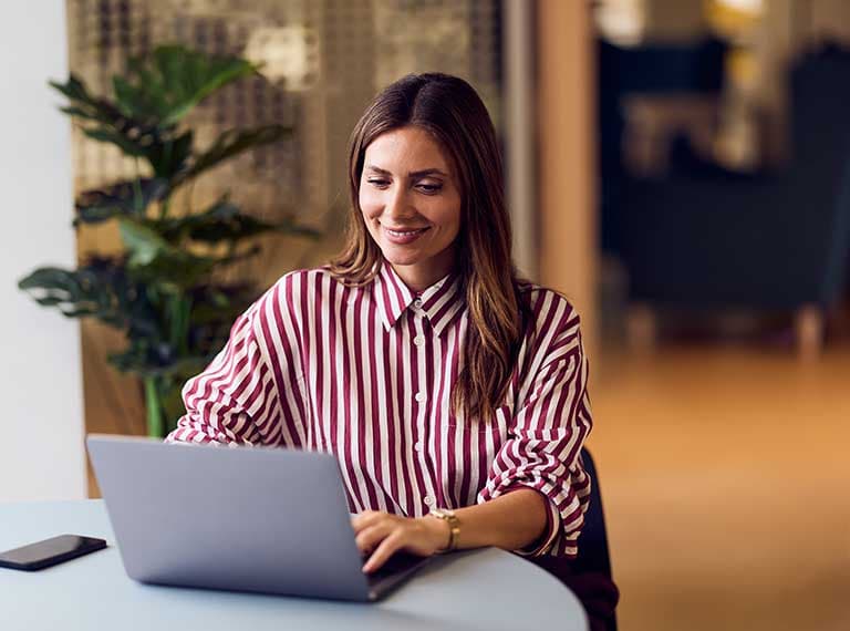 Person in red and white striped shirt smiling while working on laptop in modern office space with plants nearby.