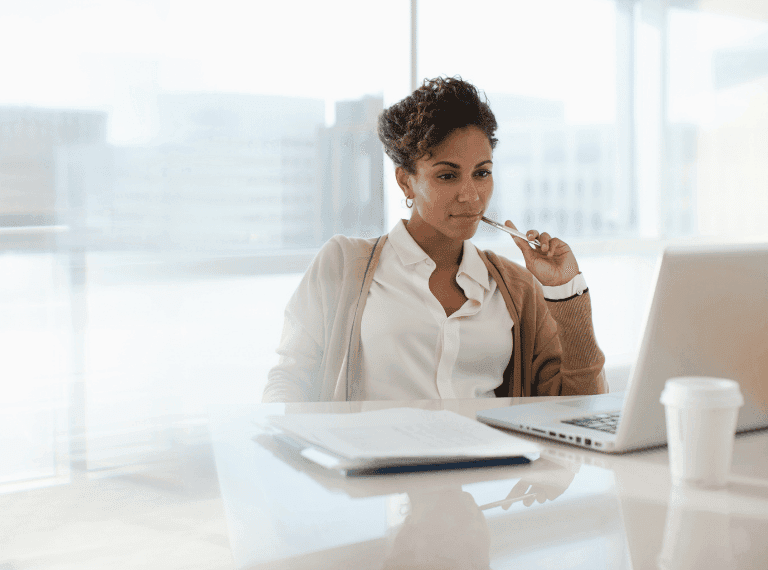 Professional woman in white shirt and beige cardigan working at laptop in bright modern office with city view