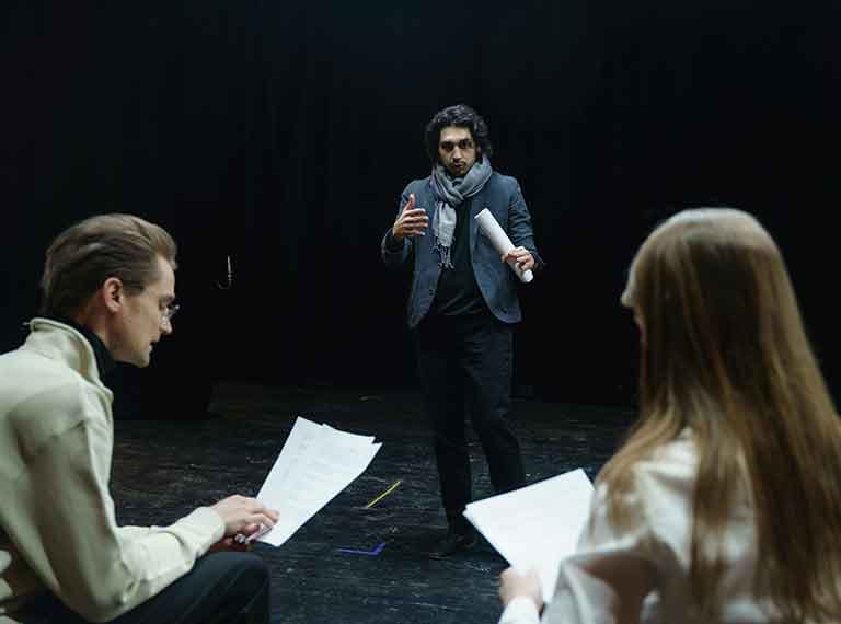 Actor in blue blazer gesturing while holding script, rehearsing with two seated people holding papers on dark stage.