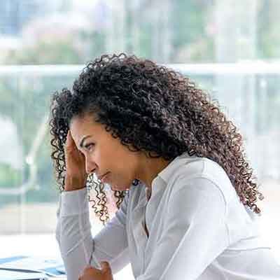 Woman with curly hair in white shirt looking stressed with hand on forehead at desk in office setting.