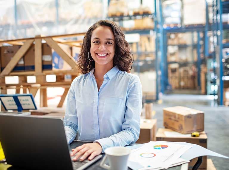Woman in light blue shirt working on laptop in warehouse with storage shelves and shipping boxes in background.