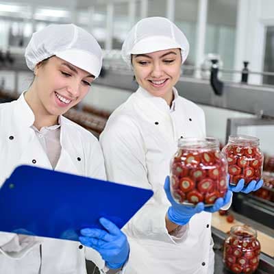 Two food production workers in white uniforms and hairnets examining jars of preserved strawberries in a factory.