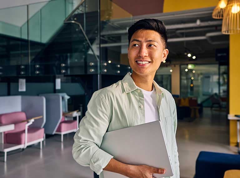 Young professional smiling in modern office space with colorful walls, holding laptop in light button-up shirt.
