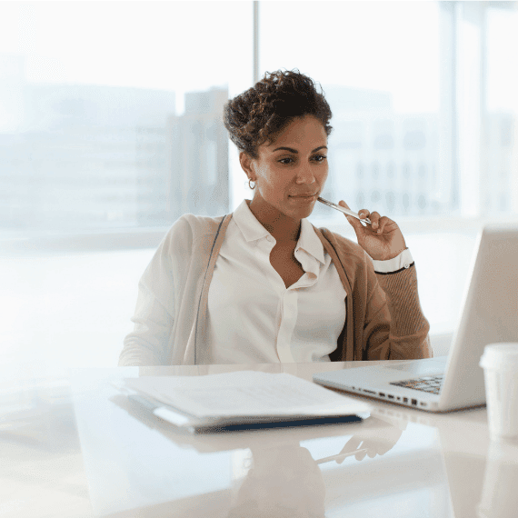 Professional woman in white shirt and beige cardigan looking at laptop while holding pen at modern office desk