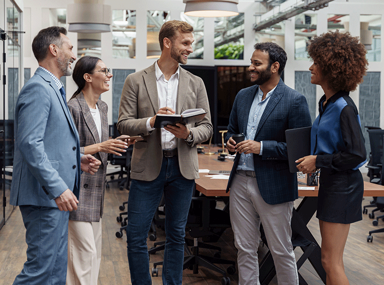 Five diverse business professionals in business casual attire having a friendly discussion in a modern office workspace.