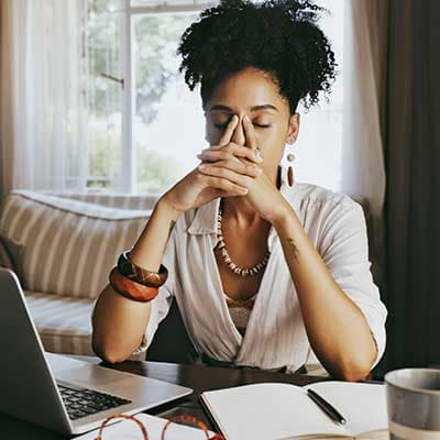 Woman with natural hair in white top sits at desk with laptop, eyes closed, hands covering nose in stressed gesture.