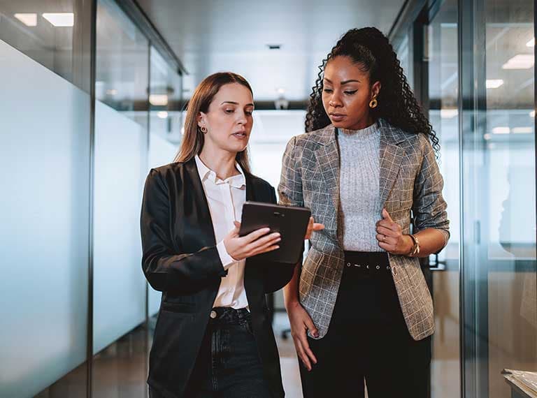 Two professionals in business attire reviewing information on a tablet in a modern office hallway with glass walls.