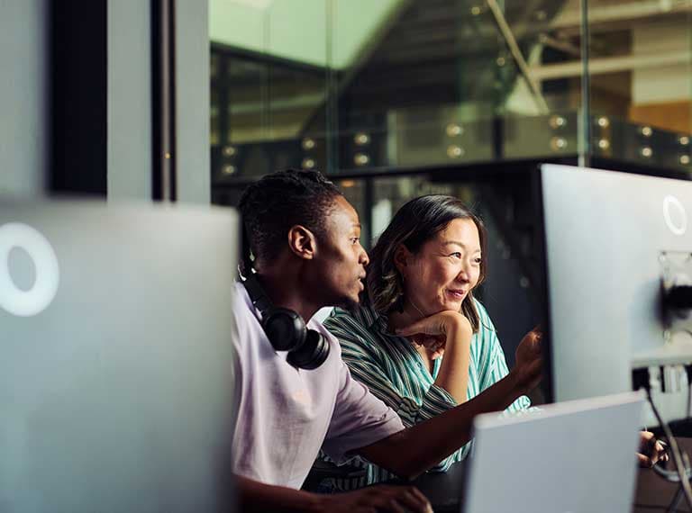 Two colleagues collaborating at a computer workstation in a modern office with glass partitions.