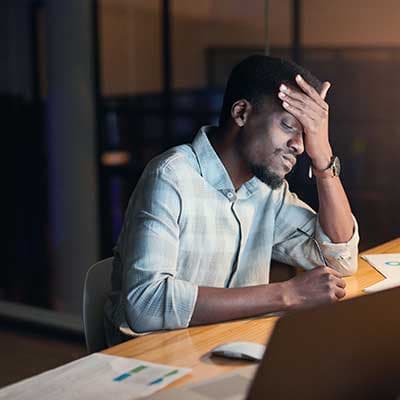 Stressed businessman in light blue shirt with hand on forehead working late at desk with documents and laptop
