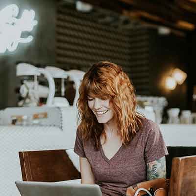 Smiling woman with red hair working on laptop in modern cafe with warm lighting and blurred background.
