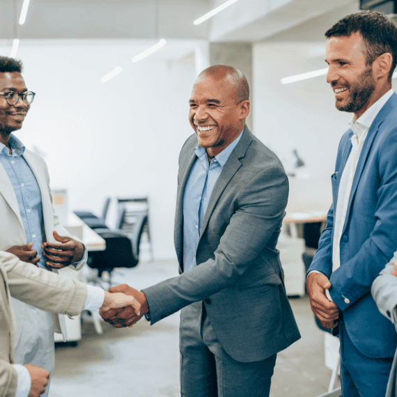 Three businessmen in suits shaking hands and smiling in a modern office setting with bright lighting.