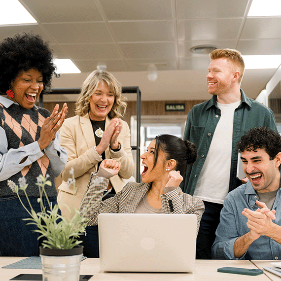 Five diverse coworkers celebrating together in an office, smiling and clapping around a laptop on a desk with a plant.